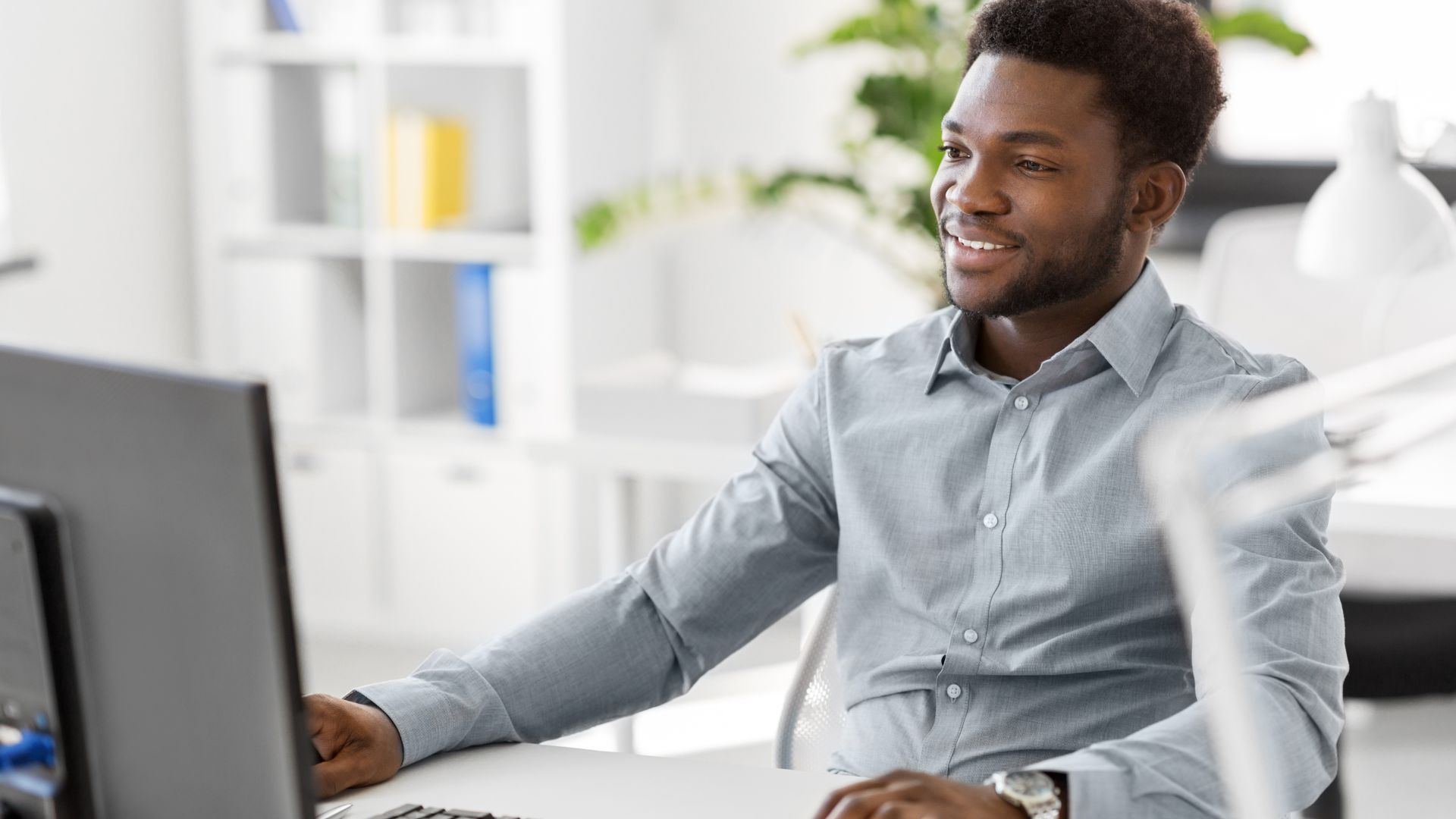 A man wearing a light grey shirt sits at a desk, smiling while looking at a computer monitor in a well-lit office space.